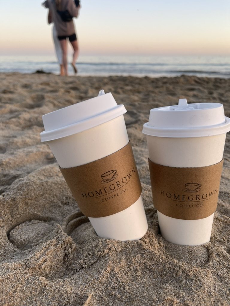 Two branded Homegrown coffee cups resting in the sand at sunset