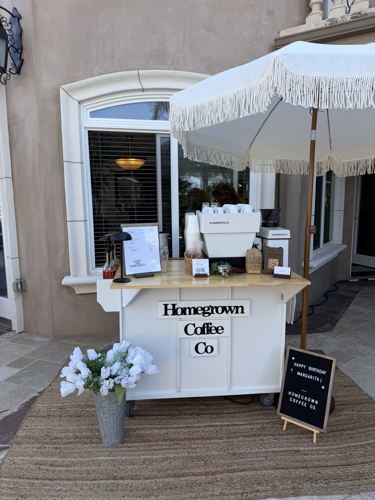 White Homegrown Coffee Co. cart beneath a fringed umbrella at an outdoor birthday celebration